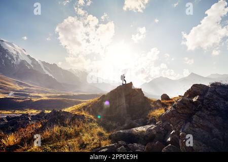 Small Man tourist with big backpack is walking climb in the green mountain valley with snowy peaks at sunset cloudy sky background Stock Photo