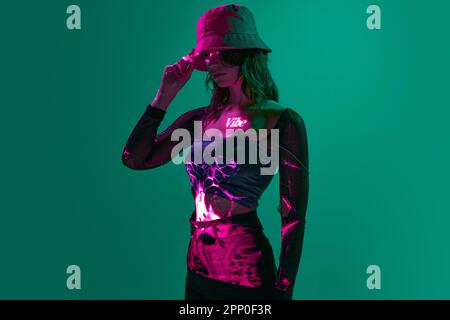 Creative portrait of young girl wearing panama with digital neon filter lights with inscryption on body on dark green mode background. Vibe Stock Photo