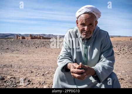 Berber old man sitting on a stone on the desert floor next to the ruins of a kasbah. Stock Photo