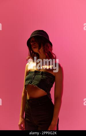Creative portrait of young girl wearing panama with digital neon filter lights with inscryption on body over pink mode background. Cringe Stock Photo