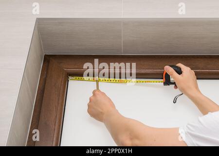 View of a man measuring a brown plastic window with a tape measure, mounting a blind on a brown plastic window Stock Photo