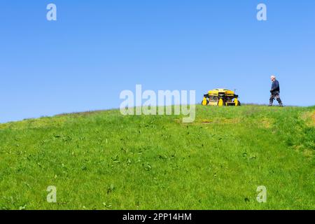 Robot mower or Robotic lawn mower mowing grass on a steep slope - man controlling a robot lawn mower - remote control industrial grass cutter uk Stock Photo