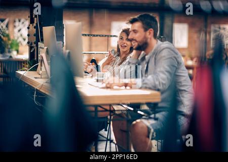 Two young office workers in casual clothes chatting on coffee break, sitting in front of desktop computers in a creative office. Business, students, e Stock Photo