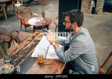 Young man sitting alone in cafe, using smarphone and listening on earphones. Nice modern cafe interior. Stock Photo
