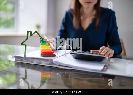 Close-up Of House Model Showing Energy Efficiency Rate In Front Of Businessperson's Hand Using Calculator Stock Photo