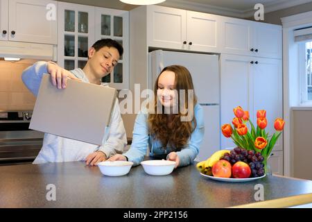 Smiling, happy boy and girl pouring frosted chocolate balls out of the cereals box for healthy cereals breakfast in morning at home pours cornflakes from a paper box into a white plate. Stock Photo