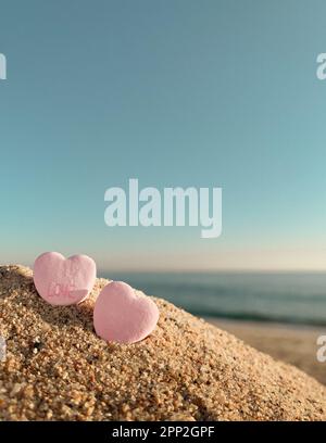 Close up low angle view of two pink candy hearts in sand on beach with ocean and blue sky in the background Stock Photo