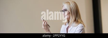 Busy hours. Stylish mature blonde woman in glasses and business attire smiling and talking on the phone, standing indoors Stock Photo