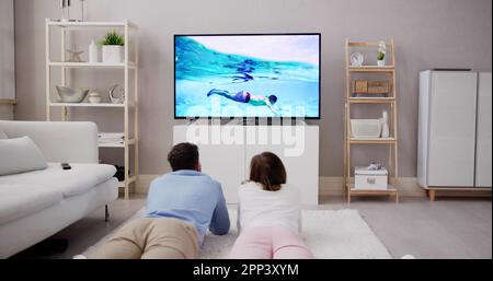 Young Happy Couple Lying On Carpet Watching Movie On Television At Home Stock Photo