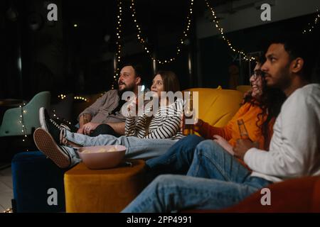 A company of multiracial friends watching a movie sitting on soft bean bags and drinking beer. Stock Photo