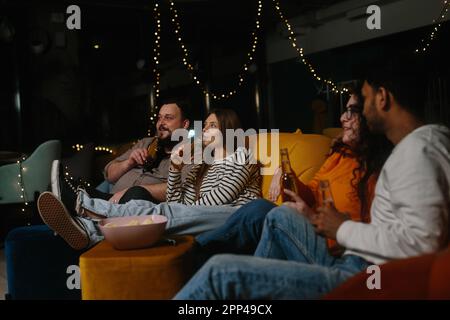 A company of multiracial friends watching a movie sitting on soft bean bags and drinking beer. Stock Photo