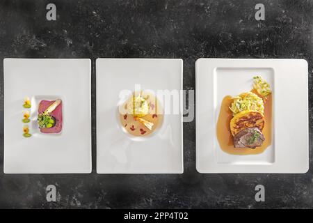 Three menu dishes on white plates, asparagus with beetroot sauce, saffron risotto with prawn, roast deer with dumpling and vegetable, dark background, Stock Photo