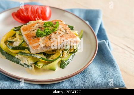 Salmon fillet with parsley garnish, zucchini vegetables and tomato on a light plate, blue napkin and a rustic wooden table, healthy low carb meal, cop Stock Photo