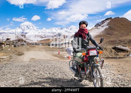 Tibetan man riding a motorcycle near  Tagong town, in the background the snowy mountain 'Yala' - Sichuan, China. Stock Photo