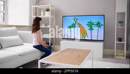 Happy Girl Sitting On Sofa Watching Cartoon On Television Stock Photo