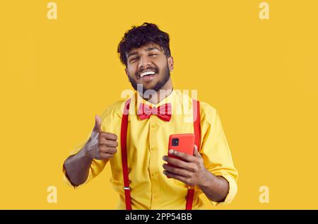 Young positive South Asian man holding mobile phone and showing thumbs up standing in yellow studio Stock Photo