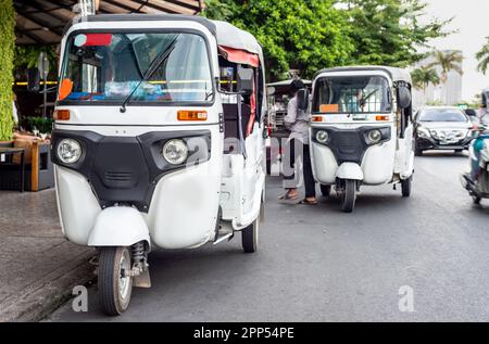 Three wheeled shiny white vehicles,with rear seat and one driver,with covered roof,waiting by the roadside in capital city of Cambodia,a popular,cheap Stock Photo