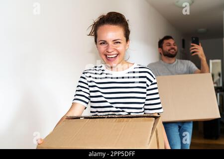 Happy young couple moving into a new apartment, carrying cardboard boxes in hands, laughing and taking first pictures of new home using phone. Stock Photo