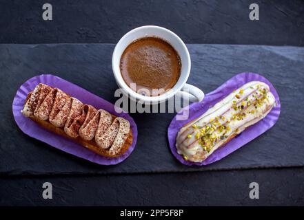 Cup of coffee and delicious eclair cakes - tiramisu and pistachio flavoured covered with cream, icing and chopped nuts, on a black background. Stock Photo