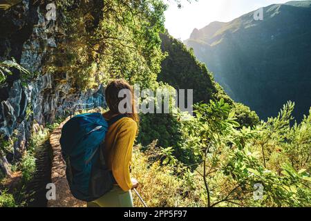 Description: Tourist woman enjoying view from below large rock wall along water channel at steep cliff through Madeira's rainforest. Levada of Caldeir Stock Photo