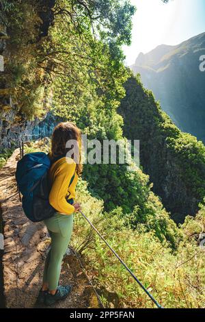Description: Tourist woman enjoying view from below large rock wall along water channel at steep cliff through Madeira's rainforest. Levada of Caldeir Stock Photo