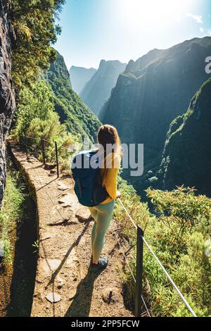 Description: Backpacker woman enjoying scenic view from below large rock wall along water channel at steep cliff through Madeira's rainforest. Levada Stock Photo