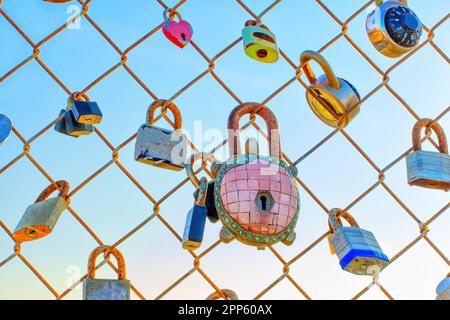 Close-up view of several padlocks hanging on a chain link fence against a backdrop of a beautiful soft sunset light. Love, commitment and devotion con Stock Photo