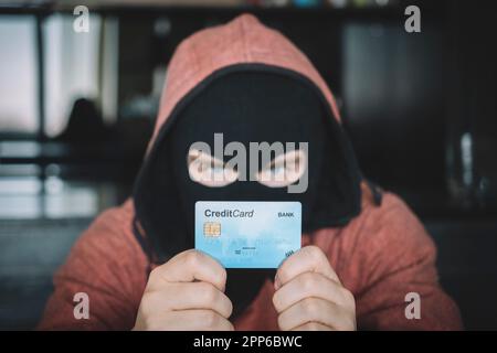 Skilled masked criminal using a stolen credit card to buy things online. A thief in a black mask holds a stolen bank card in his hands. Stock Photo