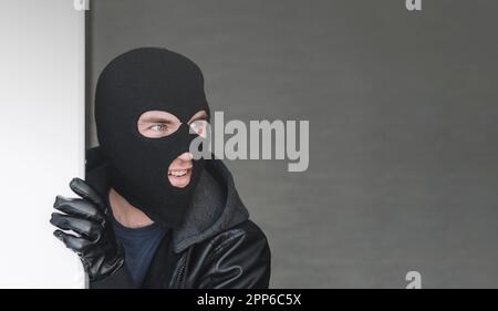 An evil thief in a mask looks out from behind a white board. Space for text and design. copy space. the surprised bandit looks away Stock Photo