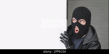 An evil thief in a mask looks out from behind a white board. Space for text and design. copy space. the surprised bandit looks away Stock Photo