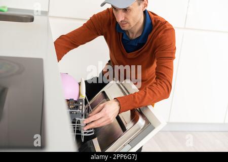 A man or a service worker in special clothes using a screwdriver attaches to the countertop or removes the dishwasher. Stock Photo