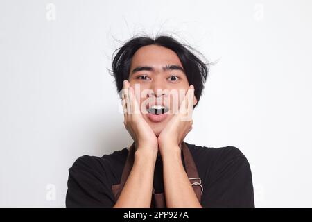 Close up portrait of shocked Asian barista man in brown apron holding hands on cheeks, feeling surprised with open mouth. Isolated image on white back Stock Photo