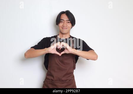 Portrait of attractive Asian barista man in brown apron speaks about own feelings, makes heart gesture over chest, expresses sympathy and love, smiles Stock Photo