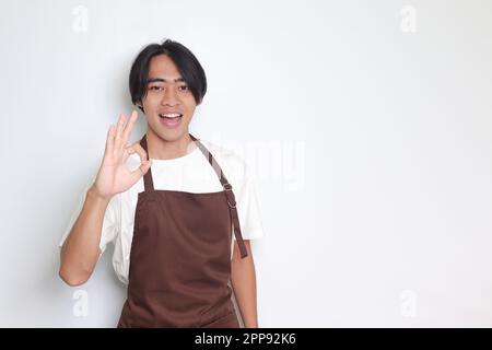 Portrait of attractive Asian barista man in brown apron showing ok hand gesture and smiling looking at camera. Advertising concept. Isolated image on Stock Photo
