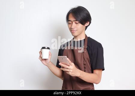 Portrait of attractive Asian barista man in brown apron holding disposable paper coffee cup while using mobile phone. Isolated image on white backgrou Stock Photo
