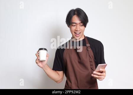 Portrait of attractive Asian barista man in brown apron holding disposable paper coffee cup while using mobile phone. Isolated image on white backgrou Stock Photo