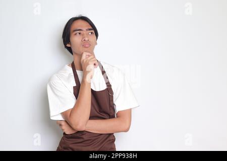 Portrait of attractive Asian barista man in brown apron thinking about question with hand on chin. Isolated image on white background Stock Photo