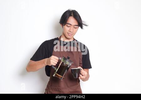 Portrait of attractive Asian barista man in brown apron pouring coffee into a cup from French press coffee maker. Isolated image on white background Stock Photo