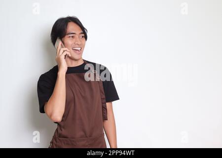 Portrait of attractive Asian barista man in brown apron having conversation while calling someone on mobile phone. Isolated image on white background Stock Photo
