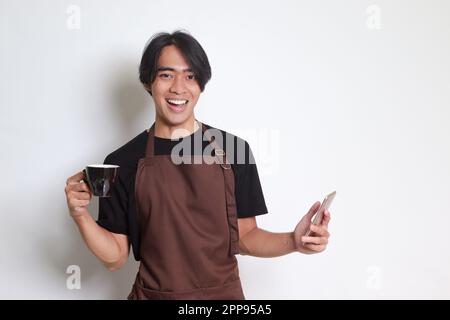 Portrait of attractive Asian barista man in brown apron holding a cup of coffee while using mobile phone and smiling at camera. Isolated image on whit Stock Photo