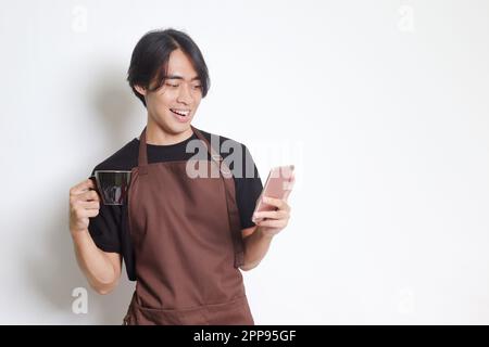Portrait of attractive Asian barista man in brown apron holding a cup of coffee while using mobile phone and smiling at camera. Isolated image on whit Stock Photo