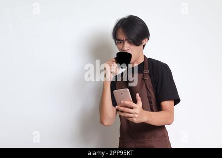 Portrait of attractive Asian barista man in brown apron drinking coffee while using mobile phone. Isolated image on white background Stock Photo
