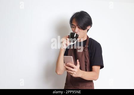 Portrait of attractive Asian barista man in brown apron drinking coffee while using mobile phone. Isolated image on white background Stock Photo