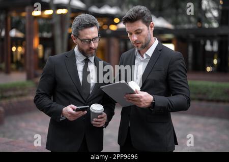 Two businessmen discussing using tablet on coffee break outdoor. Two business people talk project strategy. Two american businessmen in suits walk Stock Photo