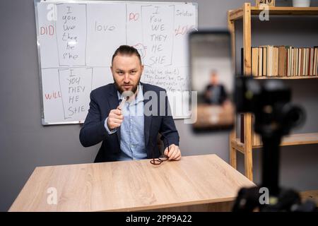 Male teacher giving online English lesson, using smartphone, pointing at webcam sitting before blackboard with grammar rules at home office. Remote Stock Photo