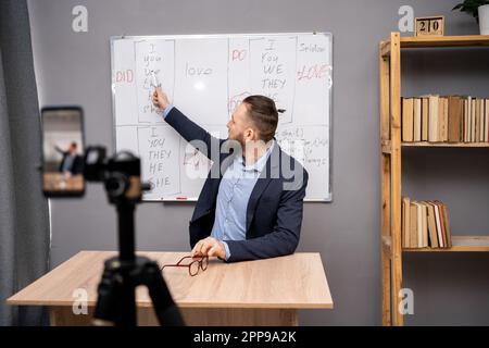 English teacher giving online lesson using whiteboard with grammar rules and smartphone, pointing at blackboard at home office. Tutor or coach engaged Stock Photo