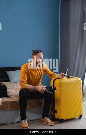 Traveler fun tourist man in casual clothes hat with suitcase using mobile cell phone booking hotel or tickets, sitting on bed at home Air flight Stock Photo