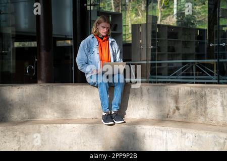 Concentrated self-sufficient man with laptop sits near building works as programmer in IT company Stock Photo