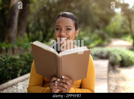 Happy brazilian lady student sitting in park outdoors, reading book, looking and smiling at camera, copy space Stock Photo