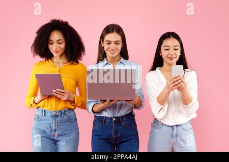 Three multiracial women using gadgets and looking at screens, browsing internet on pink studio background Stock Photo